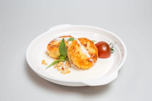 Two round fried cheese pancakes with sour cream and cherry tomatoes on a white plate on a white background. Isolated on a white background.