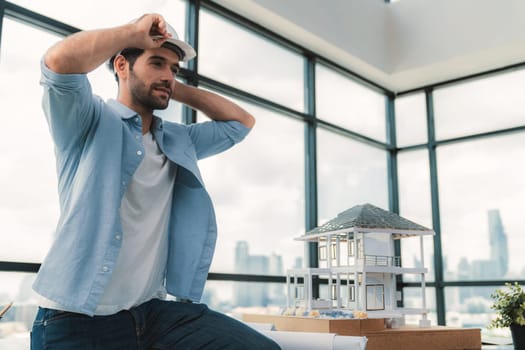 Portrait of skilled architect engineer wear safety helmet while sitting on table with house model, project plan and architectural equipment surrounded by skyscraper view. Civil engineering. Tracery.