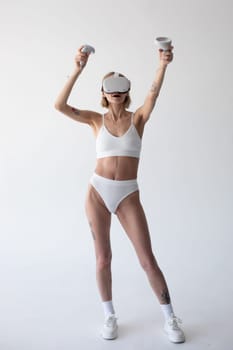 Beautiful thin girl in white shorts and a top in a virtual reality helmet posing on a white background. High quality photo