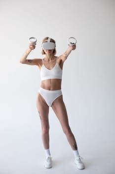 Beautiful thin girl in white shorts and a top in a virtual reality helmet posing on a white background. High quality photo