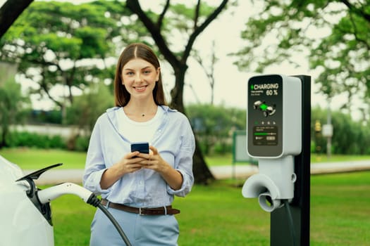 Young woman recharge EV electric vehicle battery from EV charging station and using smartphone online banking to pay for electricity in city park. Eco friendly vehicle travel with EV car. Exalt