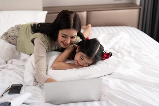 Mother and daughter lying in bed with on and smiling. Young mom working from home in bedroom with laptop.