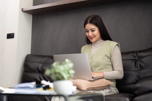 Happy young asian freelance business woman sitting on the sofa at home working on laptop computer.