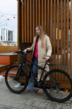 A woman is standing next to a bike parked on a city sidewalk. She appears to be checking her phone while the bike leans against a nearby fence.