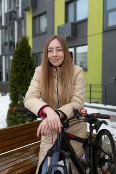 A woman clad in casual attire is seated on a wooden bench placed next to a bicycle. The woman appears relaxed as she gazes off into the distance, with the bike leaning against the bench.