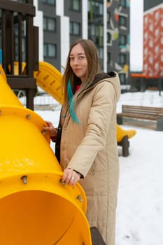 A woman is standing next to a bright yellow playground structure, looking towards the camera. The playground equipment features slides, stairs, and platforms. The scene is set in a sunny park with green grass and clear blue skies.