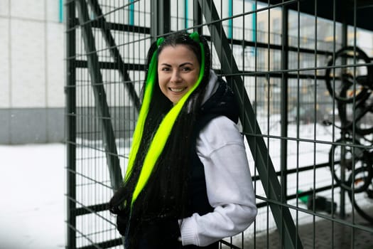 A woman wearing a green scarf is standing in front of a fence in an urban setting. She appears to be looking ahead, with a neutral expression on her face.