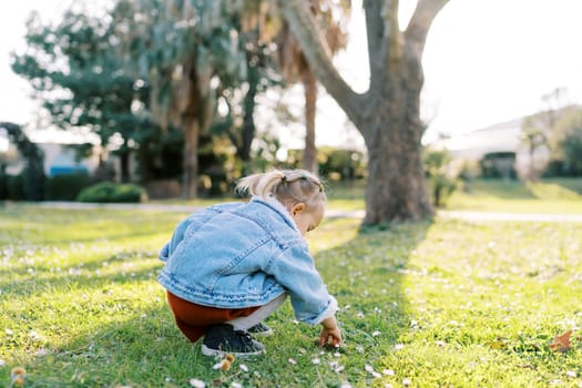 Little girl squats on a green meadow and picks the ground with her fingers. Back view. High quality photo