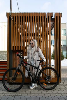 A woman is standing next to a bike in front of a wooden structure. She appears to be taking a break or exploring the area. The bike is parked on the ground, and the wooden structure provides a rustic backdrop to the scene.