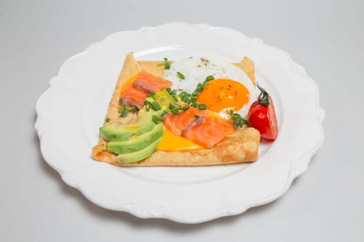 Close-up shot of a crepe with smoked salmon, avocado, egg, and tomatoes on a white plate. Square shape, elegant presentation.