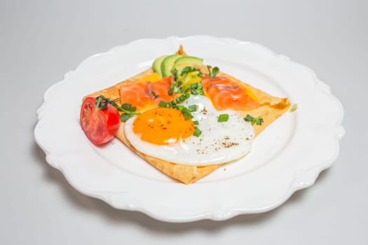 Close-up shot of a crepe with smoked salmon, avocado, egg, and tomatoes on a white plate. Square shape, elegant presentation.