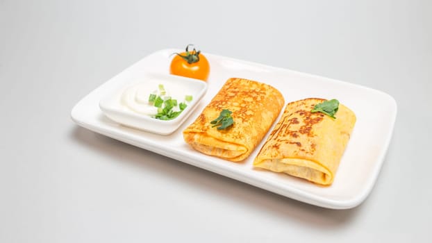 Two folded stuffed savory crepes with sour cream and chives garnish on a white plate on a white background.