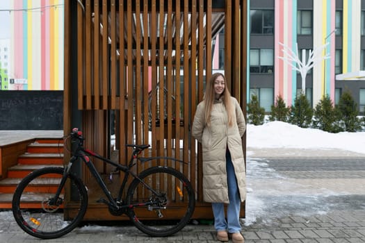 A woman in casual attire stands next to a bicycle, positioned in front of a modern building. She appears to be taking a break or checking her surroundings, with a clear sky in the background.