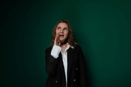 A woman is standing in front of a green wall, looking towards the camera with a neutral expression. She is wearing casual clothing and appears to be in an urban environment.