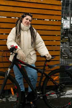 A woman with dreadlocks is standing next to a bike, appearing ready for a ride. She is wearing casual clothing and looking towards the distance.