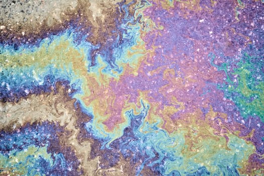 Oil residues on wet asphalt interact with sunlight, displaying a rainbow-like spectrum of colors.