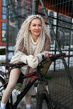 A woman is sitting on top of a black bicycle, positioned next to a wooden fence. The woman appears relaxed as she balances herself on the bike, surrounded by a simple and urban background.