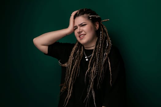 A woman with dreadlocks is standing in front of a solid green wall. She is looking directly at the camera, with a neutral expression on her face. Her hair is styled in long, thick dreadlocks that fall past her shoulders.