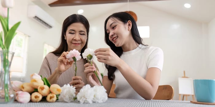 Senior mother and adult daughter happy on the table while arrange flowers in a vase together. Technology and lifestyle concept. Happy time together.