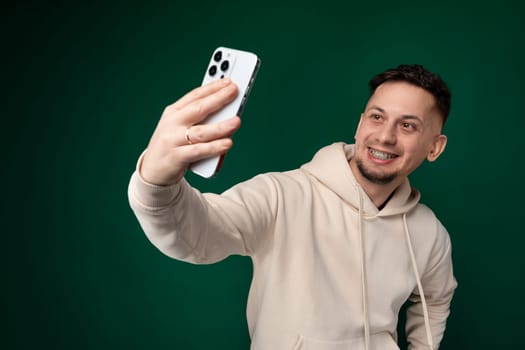 A man holding a cell phone in front of himself, smiling and posing for a selfie. The background shows a street lined with buildings and other people walking by.