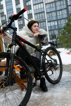 A woman is seated on a bench next to a parked bicycle in an urban setting. She looks relaxed and casual, taking a break from cycling.