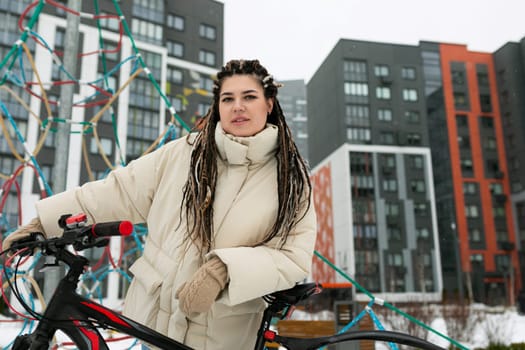A woman with dreadlocks is standing next to a bicycle on a city street. She is looking off into the distance, wearing casual clothing. The bike has a basket on the front and is parked next to a street sign.
