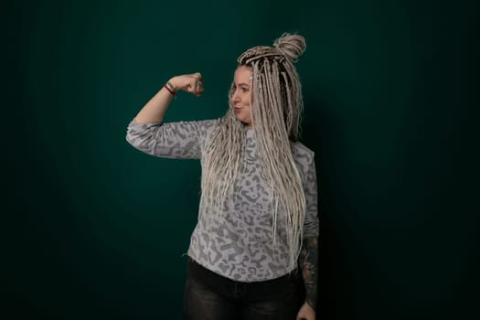 A woman with dreadlocks stands in front of a solid green wall, looking directly at the camera. Her hair is styled in long, thin locks that frame her face. She appears confident and determined, with a hint of a smile.