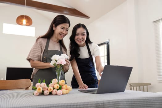 Senior mother and adult daughter happy on the table while arrange flowers in a vase together using laptop computer. lifestyle concept. Happy time together.