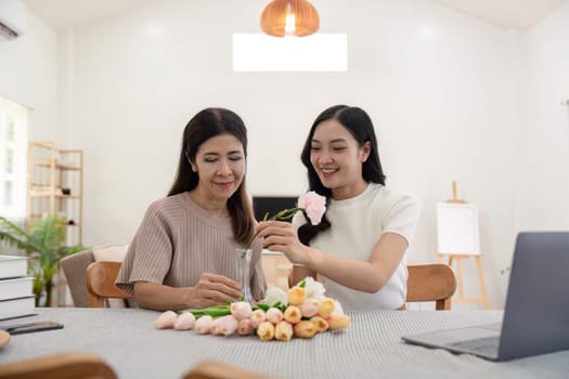 Senior mother and adult daughter happy on the table while arrange flowers in a vase together. Technology and lifestyle concept. Happy time together.