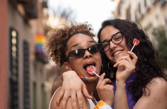 Lesbian women couple have fun while holding heart shape lollipops and smiling in Madrid city street.