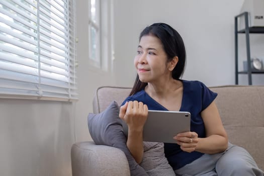 Elderly woman's leisure day watching social media on tablet, watching news on sofa in living room.