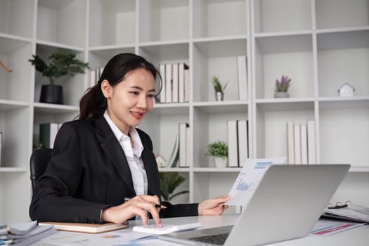 Young Asian business woman in a suit sits and reviews company work documents, checking information on a laptop on a work desk in a white office..