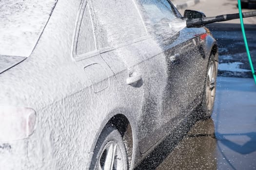 A vehicle is engulfed in soapy foam during a car wash, covering the tires, wheels, hood, and entire exterior of the car. Self-service car wash