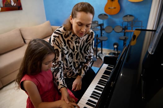 Cute little child girl sitting at grand piano near her teacher, having piano lesson at home. Different musical instruments against a blue color wall on the background. People. Education. Lifestyle