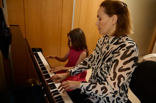 Authentic portrait of a Caucasian young adult mother and daughter sitting at grand piano, performing classic melody, toucher white and black keyboard of the piano forte, playing piano, singing song