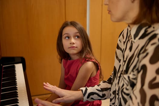Authentic portrait of beautiful Caucasian elementary age schoolkid girl in stylish red dress, smiling looking at her music teacher, sitting together at piano during individual music lesson indoors
