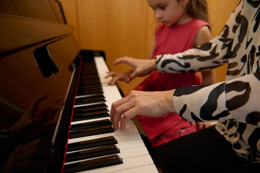 Close-up woman pianist holding hands on piano keyboard, touching black and white keys, preforming classical melody during while giving music lesson to her little student girl. People. Arts and hobbies