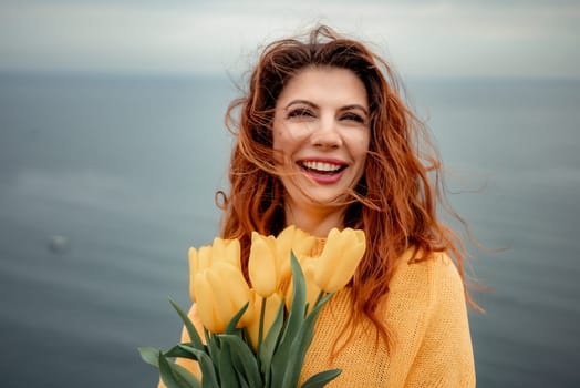 Portrait of a happy woman with hair flying in the wind against the backdrop of mountains and sea. Holding a bouquet of yellow tulips in her hands, wearing a yellow sweater.