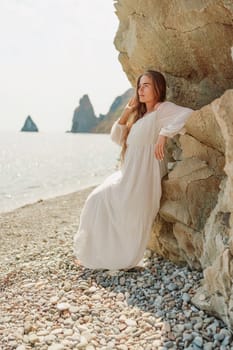 woman white dress standing on a beach, looking out at the ocean. She is lost in thought, with her hand on her hip. Concept of calm and introspection
