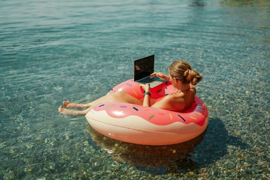 Woman laptop sea. Freelancer woman in sunglases floating on an inflatable big pink donut with a laptop in the sea. People summer vacation rest lifestyle concept