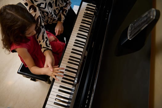 Overhead view pianist explaining to little child girl the correct position of fingers on the black and white piano keys. Caucasian cute child girl plying grand piano under the guidance of her teacher