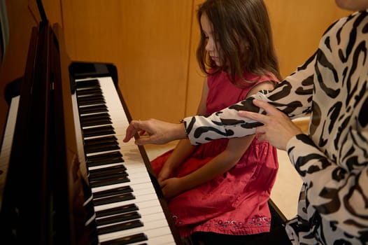Little girl in elegant red dress, taking piano lesson, passionately playing the keys under her teacher's guidance, feeling the rhythm of music. Musical education and talent development in progress