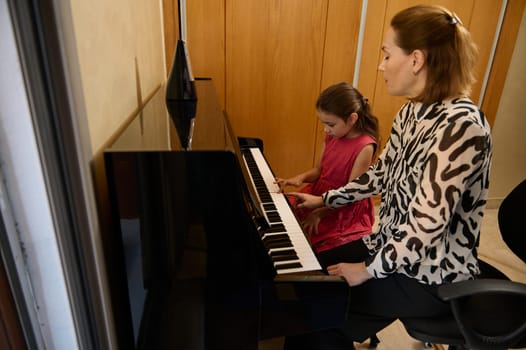 Authentic female pianist, music teacher giving individual piano lesson to her little student, sitting together at grand piano in the music class. Musical education and talent development in progress