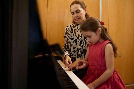 Adorable little girl in red dress, taking piano lesson, passionately playing the keys under her teacher's guidance, feeling the rhythm of melody. Musical education and talent development in progress