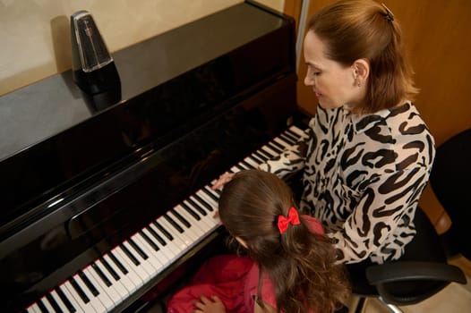 Rear view from above of a little child girl and music teacher playing together the grand piano during individual music lesson at home. Female pianist musician explaining piano lesson.