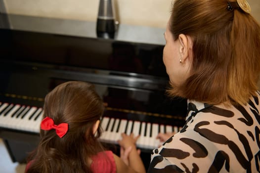 Rear view of a female musician pianist giving a piano lesson to a little schoolgirl during individual music class. Education, kids entertainment and artistic development. Hobbies and leisure