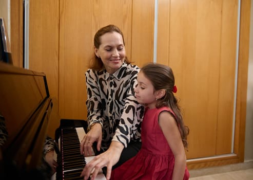Loving mother and daughter pianists playing piano together, performing a melody for Christmas during music lesson together. Cute baby girl in elegant red dress, listens to her mom playing pianoforte