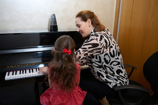 Inspired happy woman pianist, music teacher performing melody on piano forte, explaining piano lesson to a little girl, sitting nearby on a stool. Musical education and talent development in progress