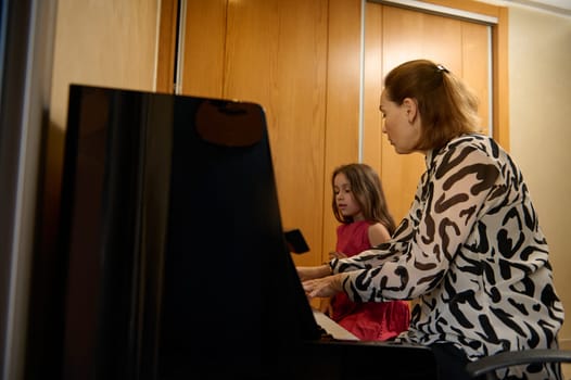 Authentic side portrait of a Caucasian young adult woman, music teacher with the pupil, sitting at vintage wooden piano at home, giving a music lesson. Mother and daughter playing piano together