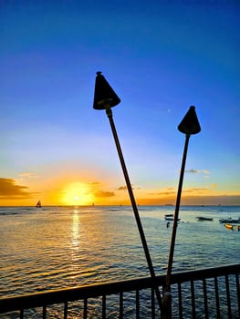 A breathtaking view of the sunset off Waikiki, where the golden hues of the setting sun gracefully merge with the tranquil blues of the ocean. Silhouetted tiki torches and a railing in the foreground add a touch of local charm, framing this serene moment where nature and culture intertwine.